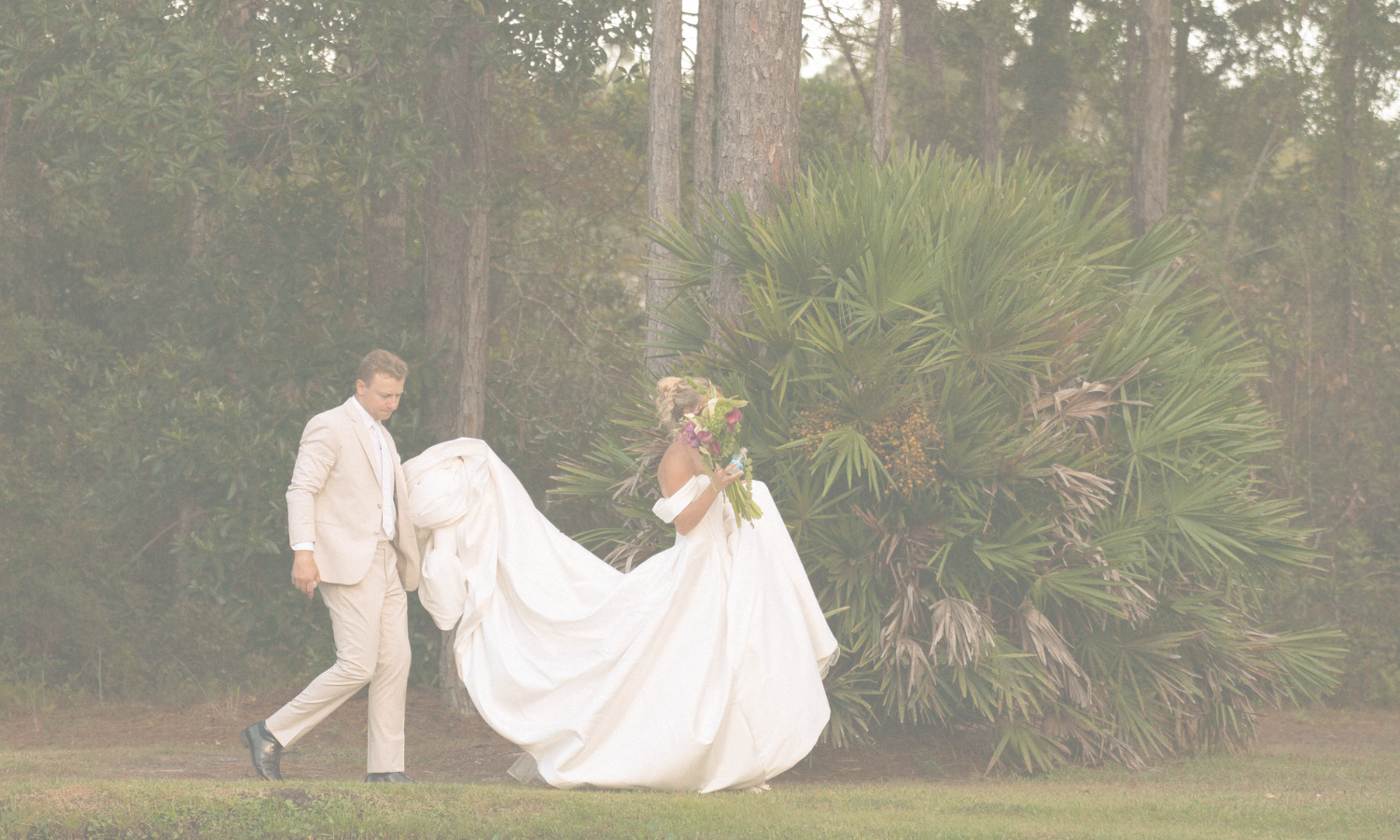 A bride in a white gown holding a bouquet, walking in a wooded area with her partner