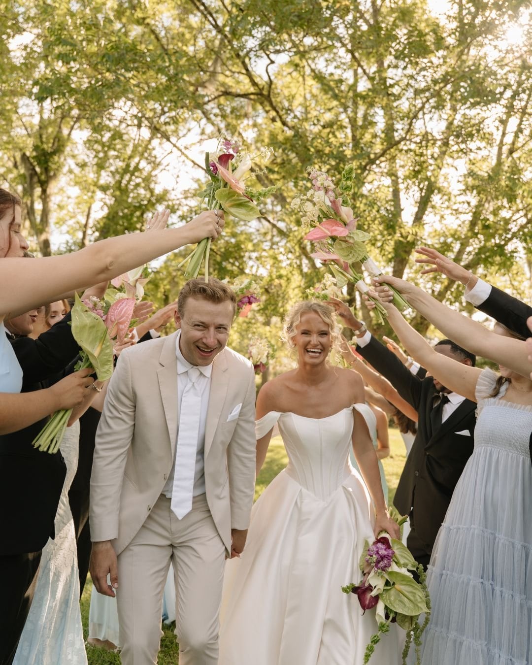 Bride and groom walking under a canopy of trees as friends raise bouquets above their heads