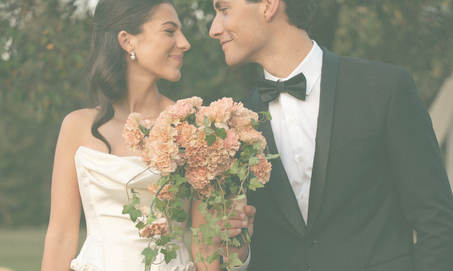 A bride and groom in wedding attire smiling at each other outdoors, the bride holding a bouquet of pink and peach flowers
